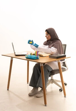 Studies and experiments in an advanced practical environment, conducting chemical reactions and development and research, a portrait of a Saudi Gulf Arab female student wearing a white coat and protective glasses holding a test tube in her hand sitting in front of a laptop conducting a scientific experiment, acquiring knowledge and experience with gestures of precision and focus, white background.