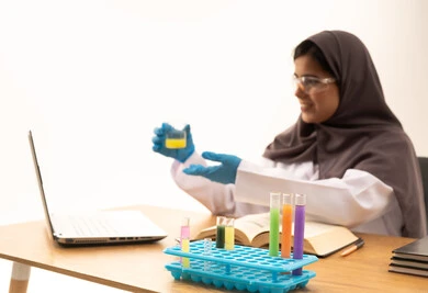 Conducting chemical reactions, development and research, studies and experiments in an advanced practical environment, a close-up portrait of a Saudi Gulf Arab female student wearing a white lab coat and protective glasses holding a test tube in her hand sitting in front of a laptop conducting a scientific experiment, acquiring knowledge and experience with gestures of precision and focus, white background.