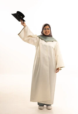 التعليم في المملكة العربية السعودية, التخرج وتحقيق الذات، بورتريه لطالبة عربية سعودية خليجية ترتدي العباءه تمسك بيدها قبعة التخرج, تعابير وجه تدل على السعاده بالتفوق والنجاح , الجسم بالطول الكامل، خلفية بيضاء