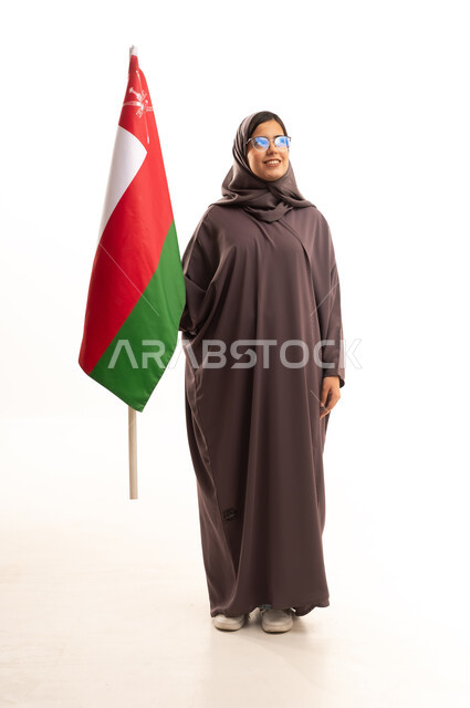 Love of the homeland as part of identity and belonging, commemorating the founding of the state in 1775 AD, cultures and national symbols, a portrait of an Arab Gulf Omani woman wearing glasses, a hijab, and an abaya holding the national flag of the country celebrating the anniversary of Omani National Day on November 18, full-body, white background.