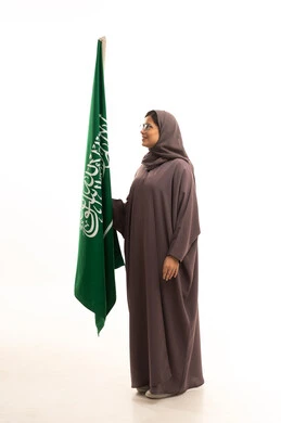 يوم العلم السعودي11 مارس، مفهوم حب الوطن و الانتماء اليه، الاحتفال بتأسيس الدولة السعودية الأولى 22 فبراير، ذكرى يوم بدينا 1727 م، بورتريه من الجانب لإمرأة عربية خليجية سعودية ترتدي العباءة و الحجاب و نظارة نظر تمسك راية وشعار المملكة تنظر بايماءات الاعتزاز و الفخر, الجسم بالطول الكامل, خلفية بيضاء
