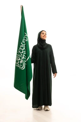 النظر الى شيء ما، الاحتفال بتأسيس الدولة السعودية الأولى 22 فبراير، مفهوم حب الوطن و الانتماء اليه، يوم العلم السعودي11 مارس، ذكرى يوم بدينا 1727 م، بورتريه لإمرأة عربية خليجية سعودية ترتدي العباءة و الحجاب تمسك راية وشعار المملكة بايماءات الاعتزاز و الفخر, الجسم بالطول الكامل, خلفية بيضاء