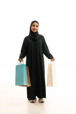 Gestures of joy after shopping and purchasing and their positive impact on women, gestures of walking and going to a specific place, seasonal discounts and offers in the markets, a portrait of a smiling Arab Saudi Gulf woman wearing a black abaya, holding colorful shopping bags in her hand, full-body, white background.