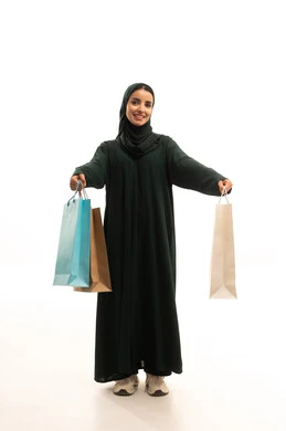 تخفيضات وعروضات موسمية في الأسواق، ايماءات السرور بعد التسوق والشراء وتأثيره الإيجابي على المرأة, بورترية لسيدة عربية سعودية خليجية مبتسمة محجبة ترتدي عباءة سوداء تحمل بيدها أكياس تسوق ملونه،  الجسم بالطول الكامل، خلفية بيضاء