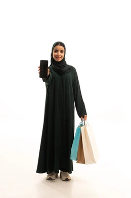 عرض شاشة سوداء فارغه عبر الجوال، متعة التسوق في إحدى المولات، استخدام التكنولوجيا والتقنية الحديثة المتطورة، بورترية لامراة عربية خليجيه سعودية ترتدي الحجاب و العباءة السوداء  تمسك بيدها الأكياس الورقية تقوم  تمسك بيدها الهاتف المحمول، الجسم بالطول الكامل، خلفية بيضاء
