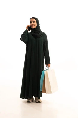 Taking advantage of seasonal offers and discounts, the joy of shopping in one of the malls, using advanced modern technology, making a phone call, a portrait of an Arab Gulf Saudi woman wearing a hijab and a black abaya holding paper bags in her hand while making a phone call via a mobile phone, full-body shot, white background.