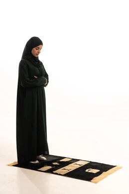 أداء الفرائض بأوقاتها،  الدعاء و التضرع لرب العباد، العبادة والتقرب إلى الله، الركوع والسجود والخشوع، مفهوم أركان الإسلام، بورتريه لشابة عربية خليجية سعودية مسلمة ترتدي الحجاب و العباءة  السوداء تقف على السجادة ، صورة الجسم بالطول الكامل، خلفية بيضاء