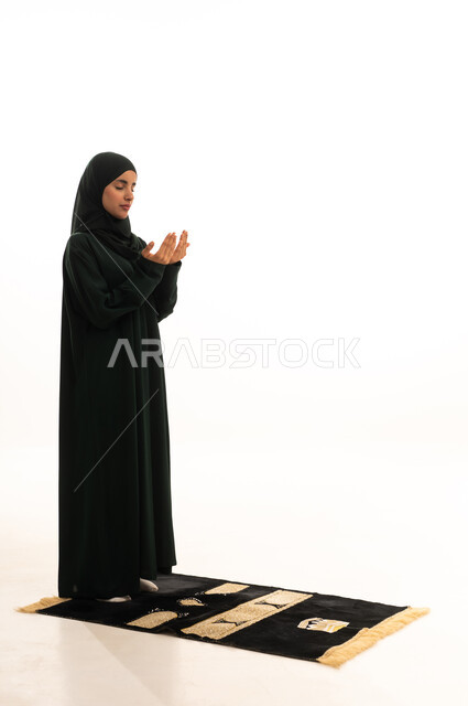 العبادة والتقرب إلى الله، الركوع والسجود والخشوع، الدعاء و التضرع لرب العباد، أداء الفرائض بأوقاتها، مفهوم أركان الإسلام، بورتريه لشابة عربية خليجية سعودية مسلمة ترتدي الحجاب و العباءة  السوداء تقف على السجادة ، صورة الجسم بالطول الكامل، خلفية بيضاء