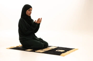 الخشوع والتضرع والطلب من الله، الاجتهاد في الطاعات والأدعية والأذكار، العبادة والتقرب الى الله، بورترية لإمرأة عربية خليجية سعودية ترتدي الحجاب والعباءة التقليدية تجلس على السجادة ترفع يديها تقوم بالدعاء، خلفية بيضاء