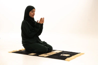 الاجتهاد في الطاعات والأدعية والأذكار، الخشوع والتضرع والطلب من الله، العبادة والتقرب الى الله، بورترية لإمرأة عربية خليجية سعودية ترتدي الحجاب والعباءة التقليدية تجلس على السجادة ترفع يديها تقوم بالدعاء، خلفية بيضاء