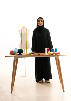 النظر الى الكاميرا بايماءات الثقه بالنفس ، مفهوم الانوثه والنعومه ،بورترية لامراه عربيه خليجيه سعوديه ترتدي العباءة السوداء والحجاب تقف امام المانيكان، ممارسة مهنة الخياطة ، الجسم بالطول الكامل، خلفية بيضاء