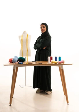 الاهتمام بالمظهر الخارجي، ايماءات الثقه بالنفس ، مفهوم الانوثه والنعومه ، بورترية لامراه عربيه خليجيه سعوديه ترتدي العباءة السوداء والحجاب تنظر إلى الكاميرا تقف بايدي متقاطعه، ممارسة مهنة الخياطة ، الجسم بالطول الكامل، خلفية بيضاء