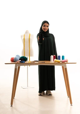 عرض شاشة سوداء فارغة، مهن و حرف يدوية , ممارسة مهنة الخياطة , بورتريه لمصممة أزياء عربية خليجية سعودية ترتدي العباءة  و الحجاب تقف أمام المانيكان تمسك بيديها الهاتف المحمول , صورة للجسم بالطول الكامل , خلفية بيضاء