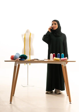 إجراء اتصال هاتفي عبر الجوال , ممارسة مهنة الخياطة , بورتريه لمصممة أزياء عربية خليجية سعودية ترتدي العباءة  و الحجاب تقف أمام المانيكان , مهن و حرف يدوية , صورة للجسم بالطول الكامل , خلفية بيضاء