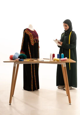 Recording measurements and notes, practicing the profession of tailoring, a portrait of an Arab Gulf Saudi fashion designer wearing an abaya and hijab standing in front of a mannequin holding a notebook and pen, mastering the craft of Saudi clothing, selecting the finest types of fabrics to make garments, using sewing tools and equipment, white background.