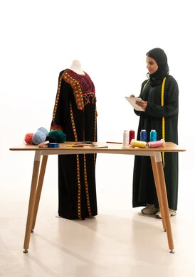 ممارسة مهنة الحياكة، تدوين القياسات والملاحظات، بورتريه لمصممة أزياء عربية خليجية سعودية ترتدي العباءة و الحجاب تقف امام المانيكان تحمل بيدها الدفتر والقلم, إتقان صناعة الملابس السعودية، اختيار أجود أنواع الأقمشة لصنع الثياب، استعمال أدوات ومعدات الخياطة، خلفية بيضاء