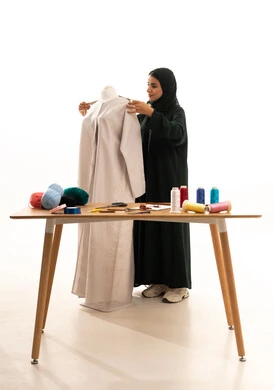 حرف و مهن يدوية في المملكة العربية السعودية، بورتريه لمصممة أزياء عربية خليجية سعودية ترتدي الحجاب و العباءة السوداء تعمل على أخذ القياسات بإستخدام الشريط المتري، ورشة عمل تصميم ازياء، مفهوم عالم الأزياء والموضة، خلفية بيضاء