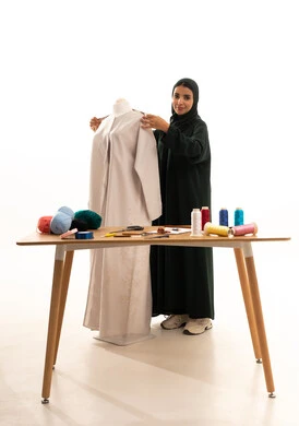 The skill of clothing design, a portrait of an Arab Gulf Saudi fashion designer wearing a hijab and a black abaya, taking measurements using a measuring tape, a fashion design workshop, the concept of the world of fashion and style, white background.