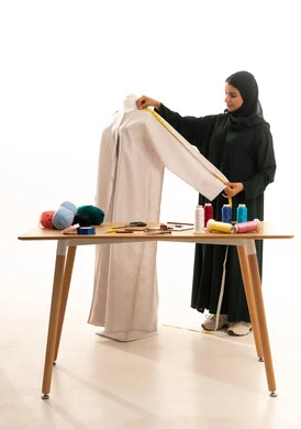 Mastering the arts of tailoring and designing women's clothing, a portrait of an Arab Gulf Saudi fashion designer wearing a hijab and a black abaya taking measurements using a measuring tape, a fashion design workshop, the concept of the fashion and style world, white background.