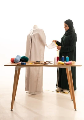 مهن وأعمال فن الخياطة، فنون ومهارات تصميم وتفصيل الملابس النسائية، بورتريه لمصممة أزياء عربية خليجية سعودية ترتدي الحجاب و العباءة السوداء تعمل على أخذ القياسات بإستخدام الشريط المتري، ورشة عمل تصميم ازياء، مفهوم عالم الأزياء والموضة، خلفية بيضاء