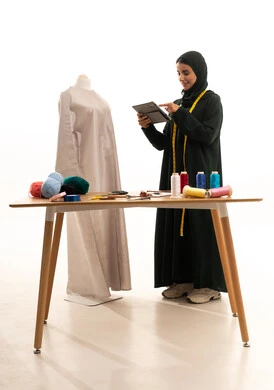 تصفح مواقع التواصل الاكتروني، إنجاز طلبات الزبائن، استخدام الجهاز اللوحي في ورشة العمل ,بورتريه لمصممة أزياء عربية خليجية سعودية ترتدي العباءة و الحجاب تمسك بيديها جهاز التابلت , محل خياطة للألبسة النسائية , الجسم بالطول الكامل، خلفية بيضاء