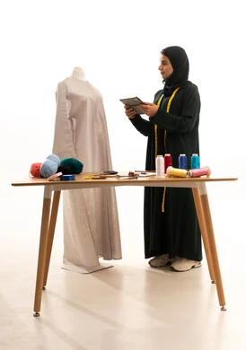 Using the tablet in the workshop, fulfilling customer orders, a portrait of a Saudi Gulf Arab fashion designer wearing an abaya and hijab looking at the mannequin holding a tablet in her hands, a women's clothing sewing shop, full-body, white background.