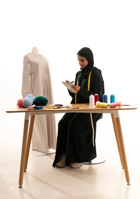 The production of women's clothing in Saudi Arabia, the practice of the tailoring profession, talent and creativity in design, a portrait of a Gulf Arab Saudi fashion designer wearing an abaya and hijab sitting in front of a mannequin holding a notebook and pen in her hands, gestures of immersion and focus, white background.