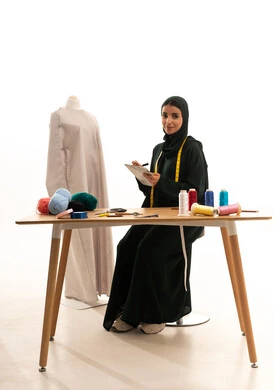ممارسة مهنة الخياطة، الموهبة و الإبداع في التصميم , بورتريه لمصممة أزياء عربية خليجية سعودية ترتدي العباءة و الحجاب تجلس امام المانيكان تمسك دفتر و قلم بيديها , النظر الى الكاميرا بايماءات السعادة و السرور، صناعة الألبسة النسائية في السعودية , خلفية بيضاء