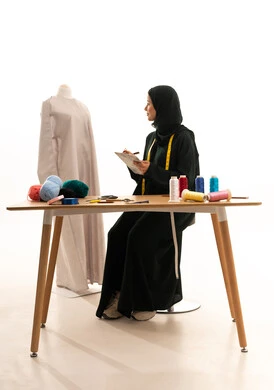 الموهبة و الإبداع في التصميم , ممارسة مهنة الخياطة , بورتريه لمصممة أزياء عربية خليجية سعودية ترتدي العباءة و الحجاب تنظر الى المانيكان تمسك دفتر و قلم بيديها , صناعة الألبسة النسائية في السعودية , خلفية بيضاء