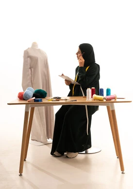 Writing daily tasks and goals, organizing and managing work, noting and recording information on paper, thinking about something, the concept of the world of fashion and style, a portrait of an Arab Gulf fashion designer from Saudi Arabia wearing a hijab and a black abaya sitting at a wooden table thinking to find solutions with a distracted expression, white background.