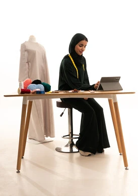 تصفح مواقع و وسائل التواصل الاجتماعي، ايماءات الاندماج و التركيز، بورترية لمصممة ازياء عربية خليجيه سعودية ترتدي الحجاب و العباءة السوداء تجلس على طاولة خشبية تقوم بانجاز مهام العمل عبر جهاز التابلت، انجاز المشاريع عن بعد، مفهوم عالم الأزياء والموضة، خلفية بيضاء