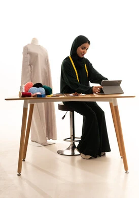 Gestures of integration and focus, browsing social media sites, a portrait of an Arab Gulf fashion designer from Saudi Arabia wearing a hijab and a black abaya sitting at a wooden table completing work tasks on a tablet, accomplishing projects remotely, the concept of the fashion and style world, white background.