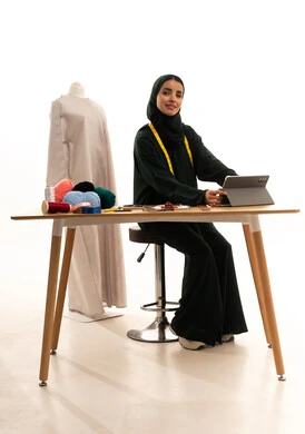 Looking at the camera with expressions of happiness, browsing social media sites, a portrait of an Arab Gulf fashion designer from Saudi Arabia wearing a hijab and a black abaya sitting at a wooden table completing work tasks on a tablet, accomplishing projects remotely, the concept of the fashion and style world, white background.