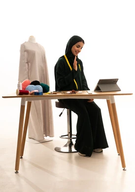 Raising the palm of the hand with gestures of greeting and peace, making video calls online, completing work tasks via tablet, managing projects remotely, playing on a mobile device, a portrait of an Arab Gulf fashion designer from Saudi Arabia wearing a hijab and a black abaya sitting at a wooden table using a tablet, the concept of the world of fashion and style, white background.