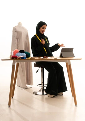 Completing work tasks via a tablet, managing projects remotely, playing on a mobile device, making video calls online, a portrait of an Arab Gulf fashion designer from Saudi Arabia wearing a hijab and a black abaya sitting at a wooden table using a tablet, the concept of the fashion and style world, white background.