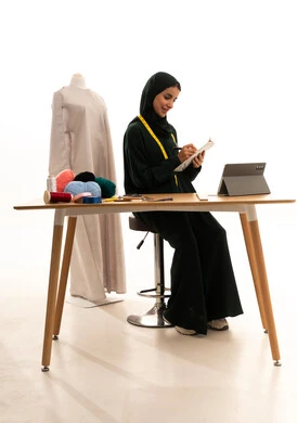 تدوين و تسجيل الملاحظات، مفهوم إدارة المشاريع عن بعد, بورتريه لإمرأة عربية خليجية سعودية محجبة ترتدي عباءة سوداء تجلس على الطاولة الخشبية تقوم بانجاز مهام العمل عبر جهاز التابلت, إستخدام اجهزة تقنية حديثة للعمل، التسوق الإلكتروني الآمن،  مفهوم عالم الأزياء والموضة، خلفية بيضاء