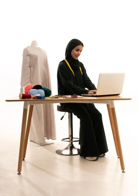 Gestures of integration and focus, working online, a portrait of an Arab Gulf Saudi fashion designer wearing a hijab and a black abaya using a laptop, the concept of the fashion and style world, white background.