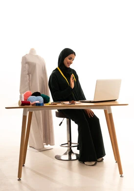 رفع كف اليد دليل على القاء التحية ورد السلام، إجراء مكالمات فيديو عبر جهاز اللابتوب، بورتريه لمصممة ازياء عربية خليجية سعودية ترتدي الحجاب و العباءة السوداء تستخدم الحاسب المحمول بايماءات السعادة والسرور، مفهوم عالم الأزياء والموضة، خلفية بيضاء