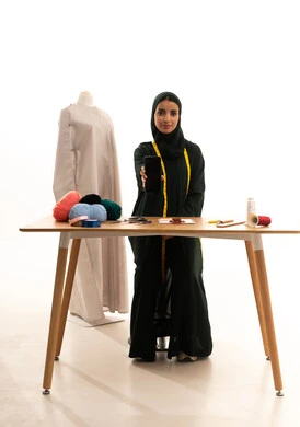 عرض تطبيقات وخدمات الهاتف المحمول، استخدام التكنولوجيا المتطورة، بورتريه لمصممة أزياء عربية خليجية سعودية ترتدي العباءة السوداء والحجاب تمسك الجوال, إتقان صناعة الملابس السعودية، اختيار أجود أنواع الأقمشة لصنع الثياب، الجسم بالطول الكامل، خلفية بيضاء