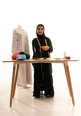 A blank black screen displayed on a mobile phone, using advanced technology, a portrait of an Arab Gulf Saudi fashion designer wearing a black abaya and hijab holding a mobile phone and pointing at it, mastering the craft of Saudi clothing, choosing the finest types of fabrics to make garments, full-body shot, white background.