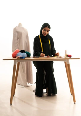 مفهوم عالم الأزياء والموضة، كتابه وتدوين الملاحظات، فنون ومهارات تصميم الملابس النسائية، بورتريه مقرب لمصممة عربية خليجية سعودية ترتدي الحجاب والعباءة تجلس على طاولة خشبية تمسك بيدها قلم تقوم بالكتابة على نوت، خلفية بيضاء