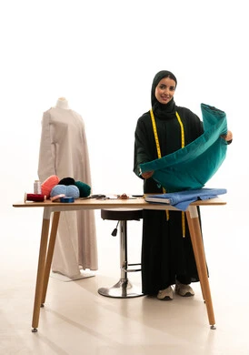 Arts and skills of women's clothing design, a portrait of a Gulf Arab Saudi designer wearing a hijab and abaya, holding fabric in her hand, looking at the camera with gestures of self-confidence, detailing modern clothing with high quality, the concept of the fashion and style world, full-length body, white background.