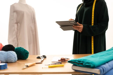 The use of modern and advanced devices and technologies, integrating work with technology, a portrait of an Arab Gulf Saudi fashion designer wearing a black abaya standing next to a mannequin and working on a tablet in a sewing workshop, designing new models, sewing equipment, and fashion sketching, white background.