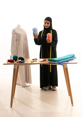 فنون ومهارات تفصيل الملابس النسائية، مفهوم عالم الأزياء والموضة، اختراع وابتكار موديلات جديدة وعصرية، أهمية المرأة في مجال الخياطة، مهن وأعمال فنية نسائية، بورتريه لامرأة عربية خليجية سعودية ترتدي عباءة تحمل بيدها بكر من خيوط الصوف، ورشة عمل تصميم ازياء، الجسم بالطول الكامل، خلفية بيضاء