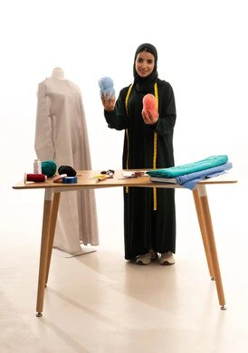 مفهوم عالم الأزياء والموضة، اختراع وابتكار موديلات جديدة وعصرية، أهمية المرأة في مجال الخياطة، مهن وأعمال فنية نسائية، بورتريه لامرأة عربية خليجية سعودية ترتدي عباءة تحمل بيدها بكر من خيوط الصوف، ورشة عمل تصميم ازياء، فنون ومهارات تفصيل الملابس النسائية، الجسم بالطول الكامل، خلفية بيضاء