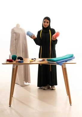اختراع وابتكار موديلات جديدة وعصرية، أهمية المرأة في مجال الخياطة، مهن وأعمال فنية نسائية، بورتريه لامرأة عربية خليجية سعودية ترتدي عباءة تحمل بيدها بكر من خيوط الصوف، ورشة عمل تصميم ازياء، فنون ومهارات تفصيل الملابس النسائية، مفهوم عالم الأزياء والموضة، الجسم بالطول الكامل، خلفية بيضاء