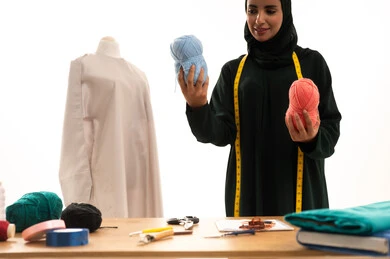 أهمية المرأة في مجال الخياطة، مهن وأعمال فنية نسائية، اختراع وابتكار موديلات جديدة وعصرية، بورتريه لامرأة عربية خليجية سعودية ترتدي عباءة تحمل بيدها بكر من خيوط الصوف، ورشة عمل تصميم ازياء، فنون ومهارات تفصيل الملابس النسائية، مفهوم عالم الأزياء والموضة، خلفية بيضاء