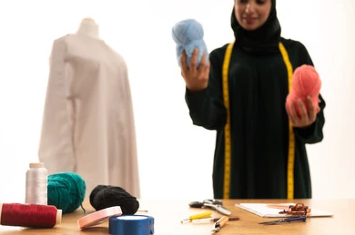 مهن وأعمال فنية نسائية، أهمية المرأة في مجال الخياطة، اختراع وابتكار موديلات جديدة وعصرية، بورتريه لامرأة عربية خليجية سعودية ترتدي عباءة تحمل بيدها بكر من خيوط الصوف، ورشة عمل تصميم ازياء، فنون ومهارات تفصيل الملابس النسائية، مفهوم عالم الأزياء والموضة، خلفية بيضاء