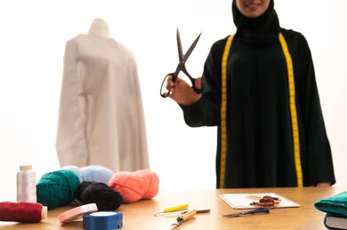 مفهوم عالم الأزياء والموضة، بورتريه لامراة عربية خليجية سعودية ترتدي العباءة السوداء و الحجاب تعمل في مهنة الخياطة تحمل بيدها مقص ، مصممة أزياء مبتكرة، ورشة عمل تصميم ازياء، مهن وأعمال فن الخياطة، فنون ومهارات تصميم وتفصيل الملابس النسائية، خلفية بيضاء
