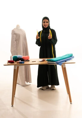 أهمية المرأة في مجال الخياطة، موديلات جديدة وعصرية، بورتريه لامرأة عربية خليجية سعودية ترتدي العباءة تحمل بيدها ابرة الكروشية، ورشة عمل تصميم ازياء، مهن وأعمال فنية نسائية،  مفهوم عالم الأزياء والموضة، الجسم بالطول الكامل، خلفية بيضاء